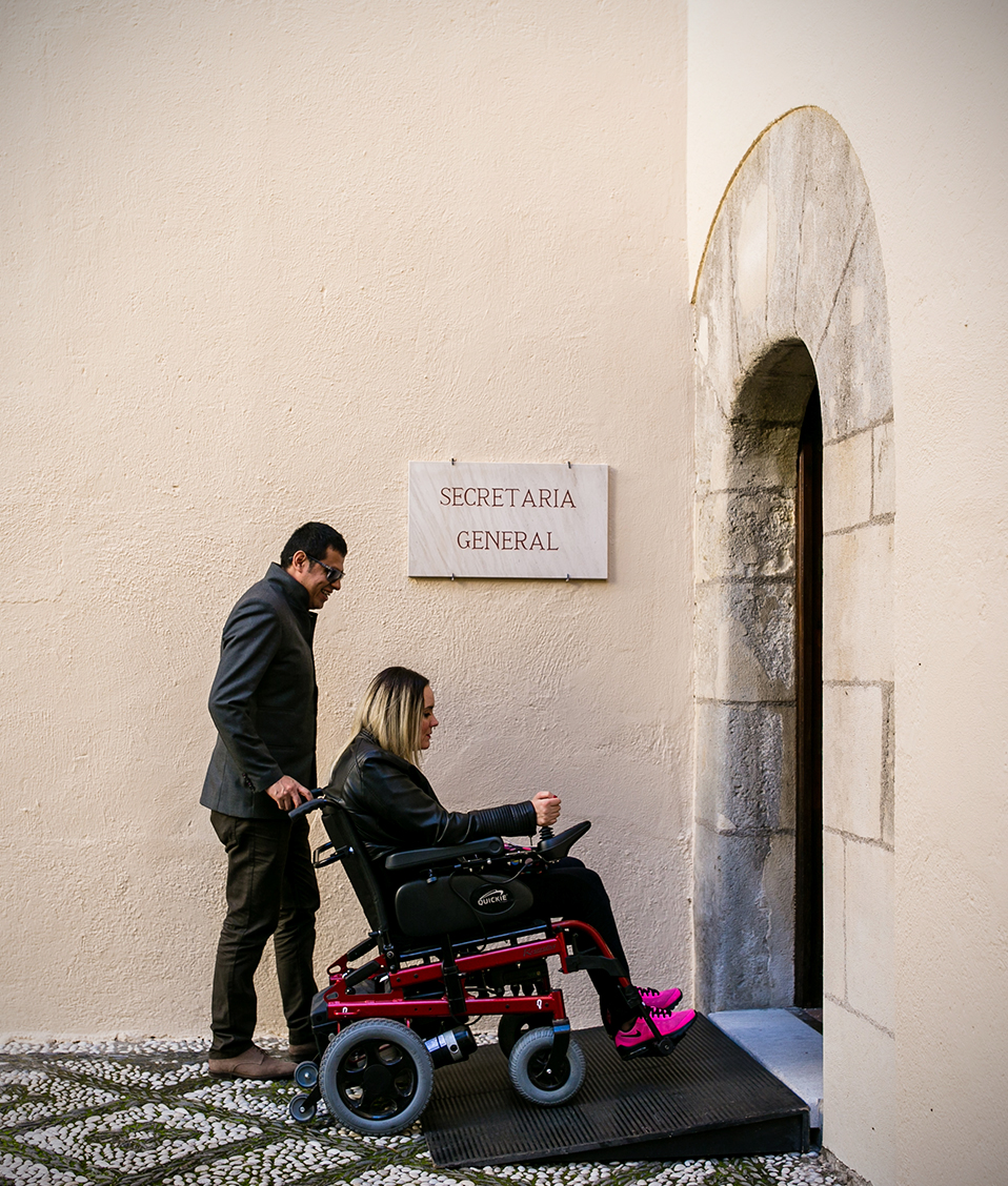 Hombre con mujer en silla de ruedas accediendo a través de una rampa de acceso a Secretaría General en patio del Hospital Real