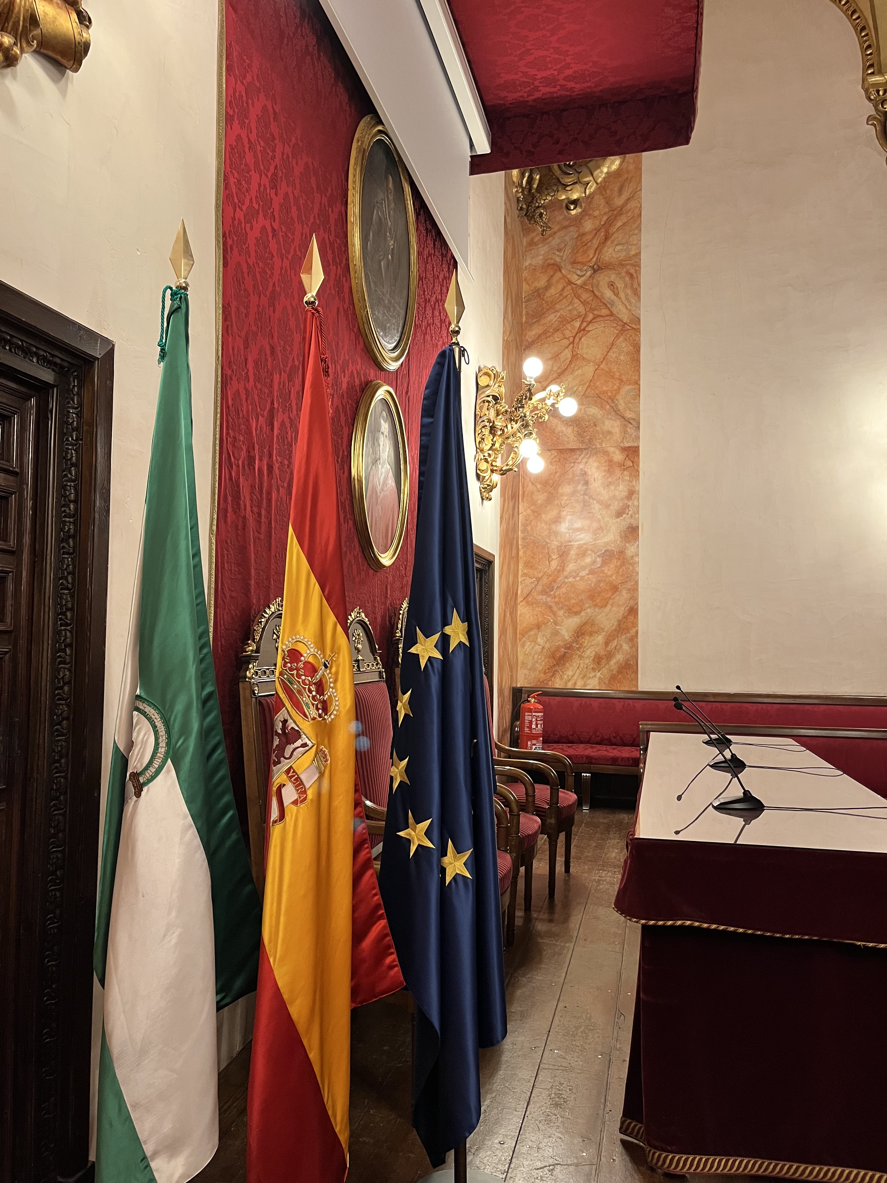 banderas de andalucía, españa y unión europea al lado de una mesa presidencial alargada con micrófonos